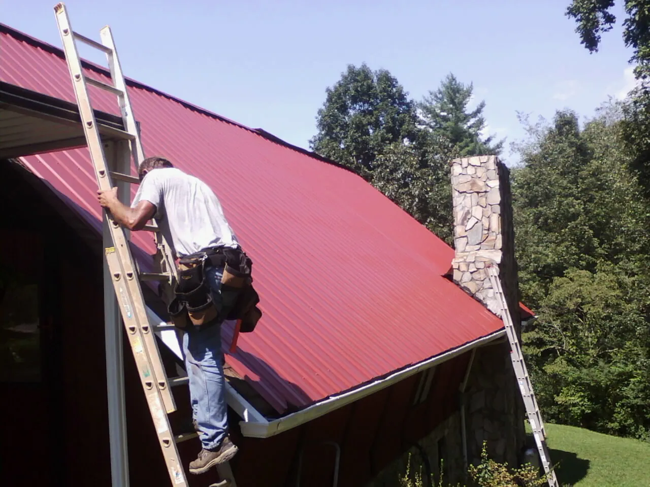 Roofer on a ladder beside a red metal roof and stone chimney — Soffit & Fascia Repair by Roofing Brothers in Plummer